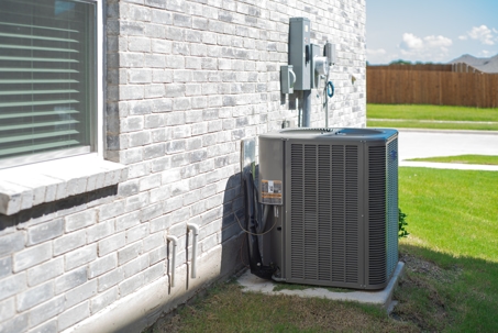 Side windows of new construction home and central air conditioning condenser unit mounted on concrete slab beside brick house wall, utility meters and electrical disconnect, suburbs